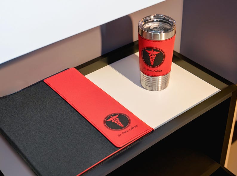K&ouml;nnte beinhalten: Ein schwarzer und roter Lederordner mit einem medizinischen Symbol und dem Text "Dr. Dara Labron" auf der Vorderseite. Eine Edelstahl-Reisebecher mit einer roten Lederh&uuml;lle und dem gleichen medizinischen Symbol und Text befindet sich auf einer wei&szlig;en Oberfl&auml;che.