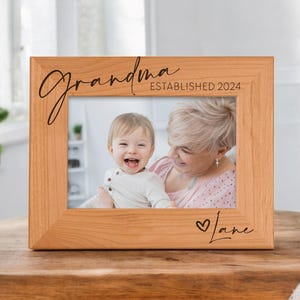 May include: A wooden picture frame with the word "Grandma" in cursive script and the text "ESTABLISHED 2024". The frame holds a photo of a smiling child and an adult. The name "Jane" is written below the photo with a heart symbol.