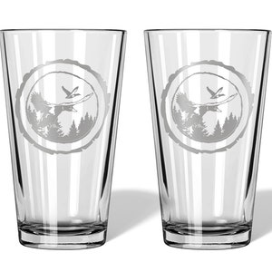 May include: Two clear glass pint glasses with an etched design of a bird flying over a forest scene inside a tree ring.