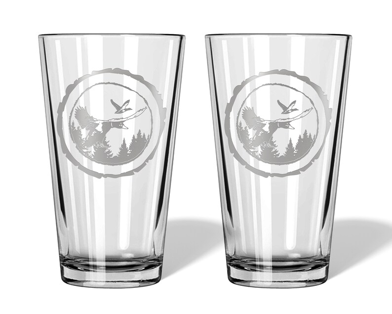 May include: Two clear glass pint glasses with an etched design of a bird flying over a forest scene inside a tree ring.