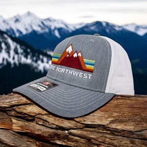 May include: Gray and white trucker hat with a red, orange, yellow, and blue mountain range graphic and the text "Pacific Northwest" embroidered on the front.