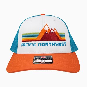 May include: White trucker hat with a blue mesh back. The front of the hat features an embroidered design of mountains and a sunset with the text "PACIFIC NORTHWEST".