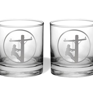 May include: A pair of clear glass tumblers, each featuring a circular etched design on the front. The engraving depicts a silhouette of a utility worker climbing a wooden power pole, complete with crossarms and electrical lines in a circular frame.