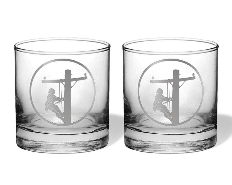 May include: A pair of clear glass tumblers, each featuring a circular etched design on the front. The engraving depicts a silhouette of a utility worker climbing a wooden power pole, complete with crossarms and electrical lines in a circular frame.