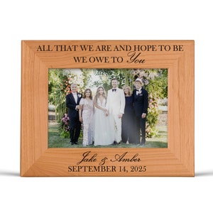 May include: A light brown wooden picture frame featuring the engraved text ALL THAT WE ARE AND HOPE TO BE WE OWE TO YOU at the top and Jake & Amber SEPTEMBER 14, 2025 at the bottom, containing a photograph of six people standing outdoors.
