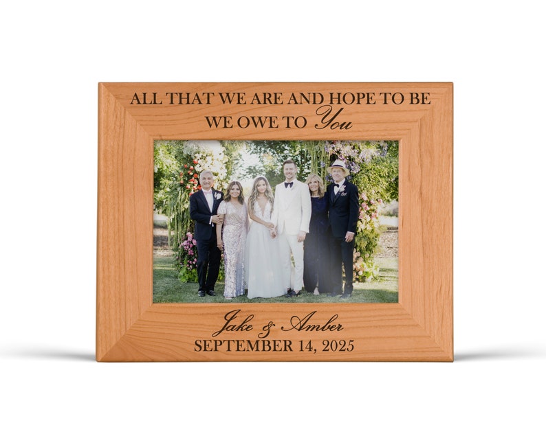 May include: A light brown wooden picture frame featuring the engraved text ALL THAT WE ARE AND HOPE TO BE WE OWE TO YOU at the top and Jake & Amber SEPTEMBER 14, 2025 at the bottom, containing a photograph of six people standing outdoors.
