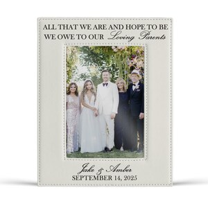 May include: A rectangular white leatherette picture frame featuring black text that reads ALL THAT WE ARE AND HOPE TO BE WE OWE TO OUR Loving Parents at the top and Jake & Amber SEPTEMBER 14, 2025 at the bottom, framing a wedding photograph.