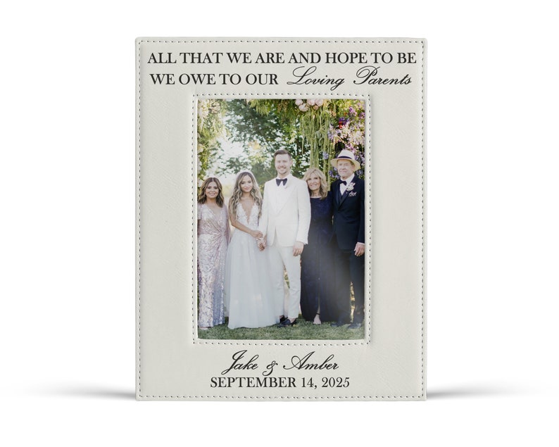 May include: A rectangular white leatherette picture frame featuring black text that reads ALL THAT WE ARE AND HOPE TO BE WE OWE TO OUR Loving Parents at the top and Jake & Amber SEPTEMBER 14, 2025 at the bottom, framing a wedding photograph.