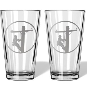 May include: A pair of clear glass pint glasses, each featuring a circular etched design. The engraving depicts a silhouette of a utility worker climbing a wooden power pole, complete with crossarms and electrical lines, centred on the glass surface.