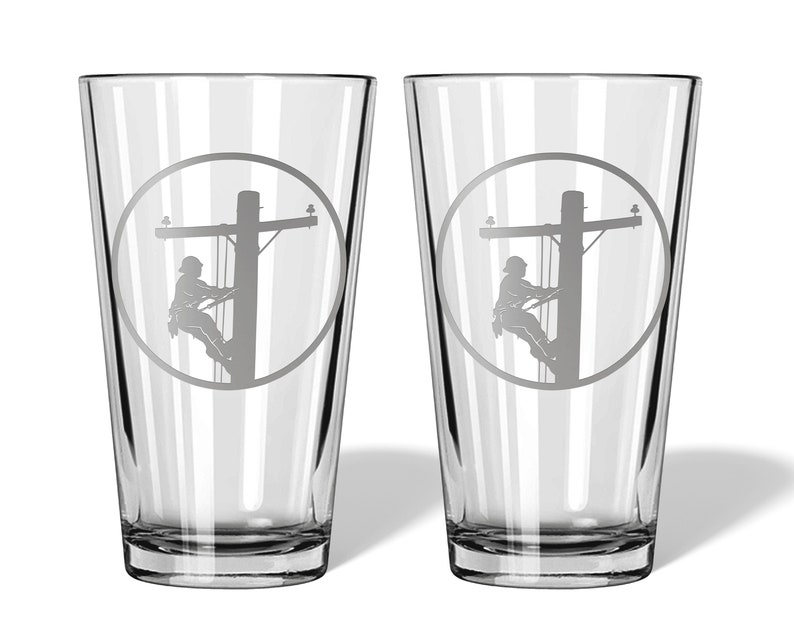 May include: A pair of clear glass pint glasses, each featuring a circular etched design. The engraving depicts a silhouette of a utility worker climbing a wooden power pole, complete with crossarms and electrical lines, centred on the glass surface.