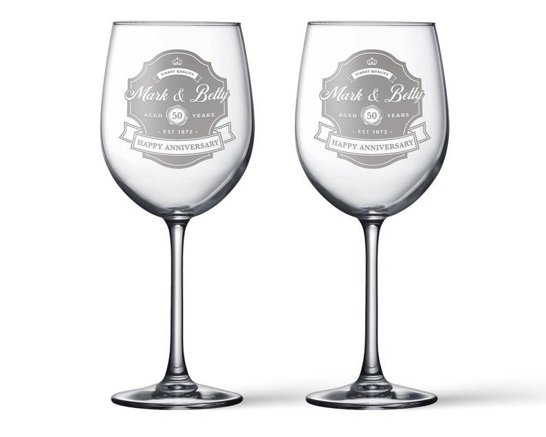 May include: Two clear glass wine glasses with etched designs. Each glass has a banner with the text "Mark & Betty" and "Happy Anniversary". The banner also includes the text "Aged 50 Years" and "Est 1972".