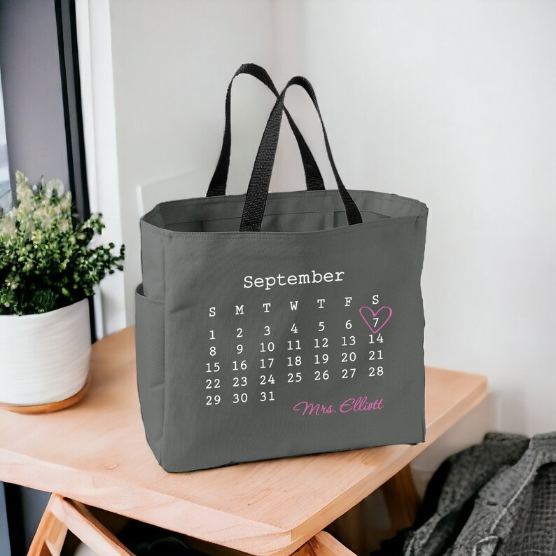 May include: A grey tote bag with black handles featuring a September calendar design. The 7th is highlighted with a pink heart. The text "Mrs. Elliott" is written in pink script below the calendar.