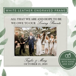 May include: A white leather picture frame with stitched edges, engraved with the text ALL THAT WE ARE AND HOPE TO BE WE OWE TO OUR Loving Parents, Taylor & Mary, and OCTOBER 21, 2023, displaying a wedding photo of a couple and guests.