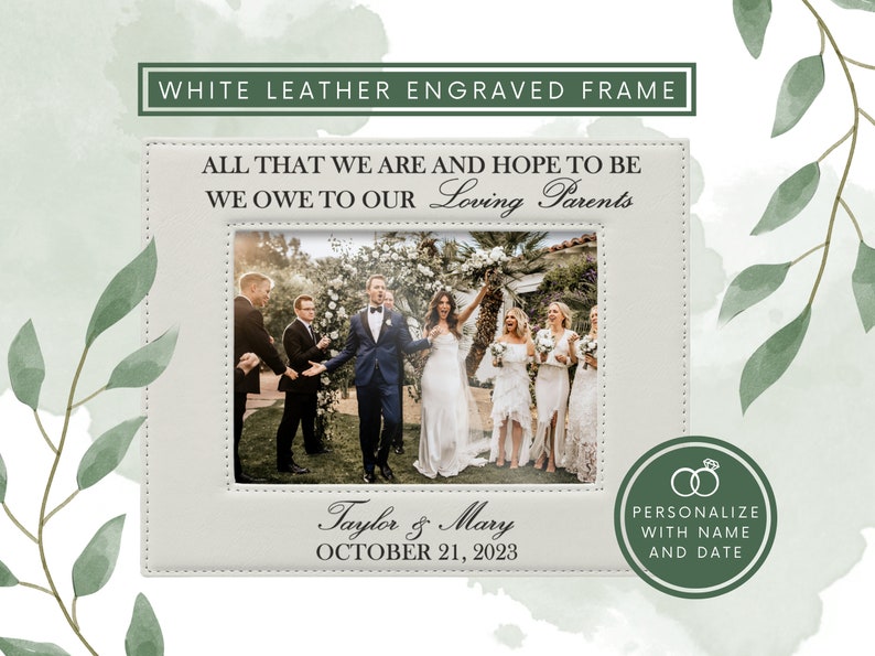 May include: A white leather picture frame with stitched edges, engraved with the text ALL THAT WE ARE AND HOPE TO BE WE OWE TO OUR Loving Parents, Taylor & Mary, and OCTOBER 21, 2023, displaying a wedding photo of a couple and guests.