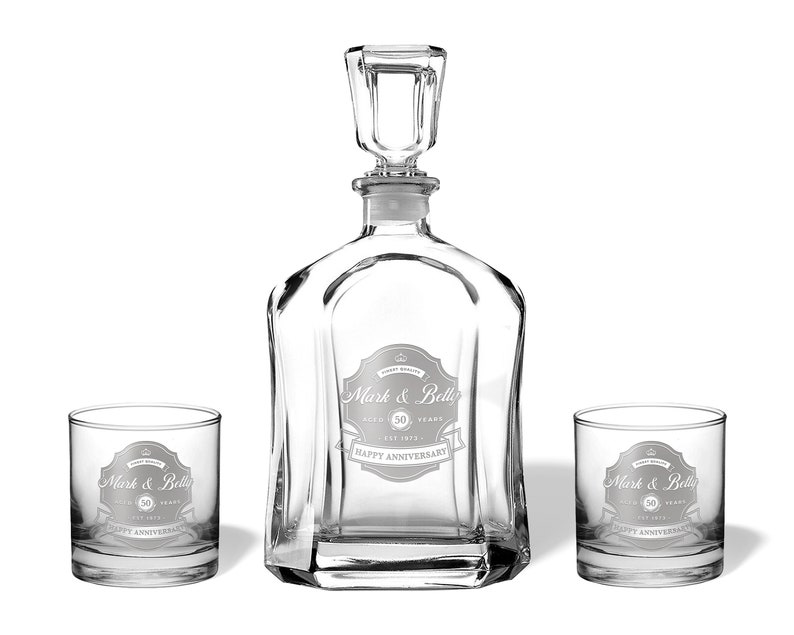 May include: A clear glass decanter with a stopper and two matching glasses. The decanter and glasses are engraved with the text "Mark & Belly 50 Years Happy Anniversary".