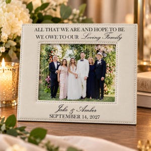 May include: A rectangular off-white leather picture frame with stitched edges. The top reads ALL THAT WE ARE AND HOPE TO BE WE OWE TO OUR Loving Family. The bottom reads Jake & Amber SEPTEMBER 14, 2027. It holds a photo of five people.