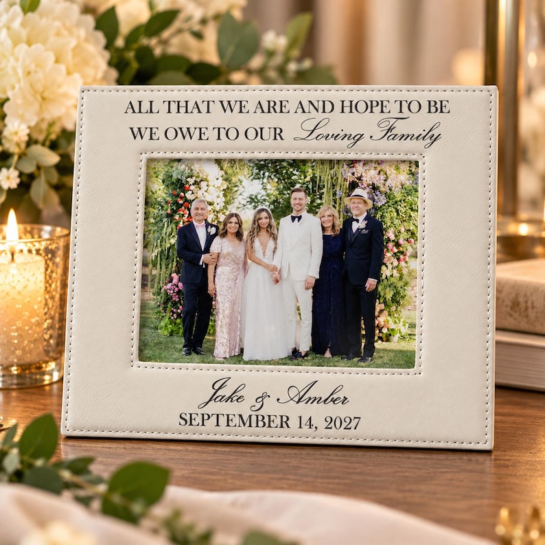May include: A rectangular off-white leather picture frame with stitched edges. The top reads ALL THAT WE ARE AND HOPE TO BE WE OWE TO OUR Loving Family. The bottom reads Jake & Amber SEPTEMBER 14, 2027. It holds a photo of five people.