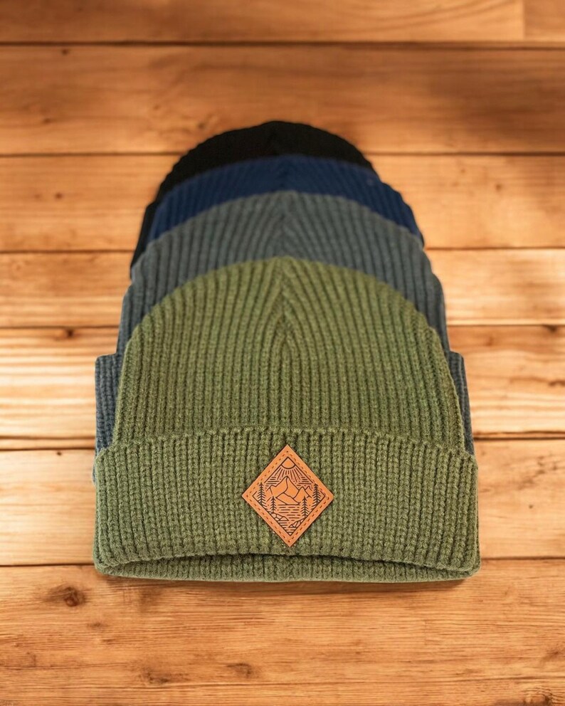 Puede incluir: Una pila de cuatro gorros de punto en diferentes tonos de azul y verde. El gorro superior es verde y presenta un parche de cuero marr&oacute;n con un dise&ntilde;o de escena de monta&ntilde;a.