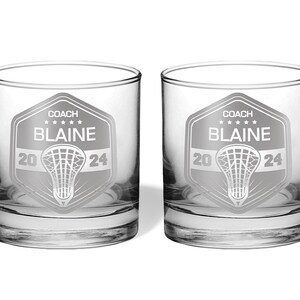 May include: Two clear, round-bottomed glasses with a lacrosse-themed design. Each glass features a shield with the word "COACH", the name "BLAINE", and the year "2024". A lacrosse net graphic is centered below the name. The glasses are ideal for beverages.