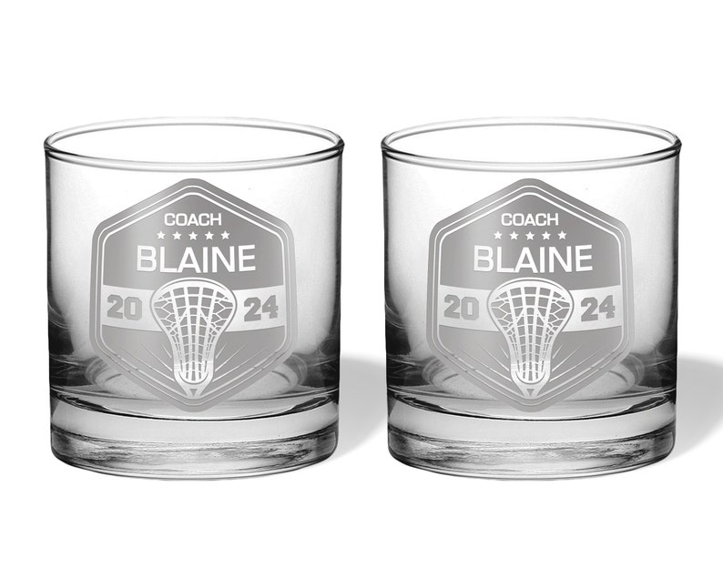 May include: Two clear, round-bottomed glasses with a lacrosse-themed design. Each glass features a shield with the word "COACH", the name "BLAINE", and the year "2024". A lacrosse net graphic is centered below the name. The glasses are ideal for beverages.
