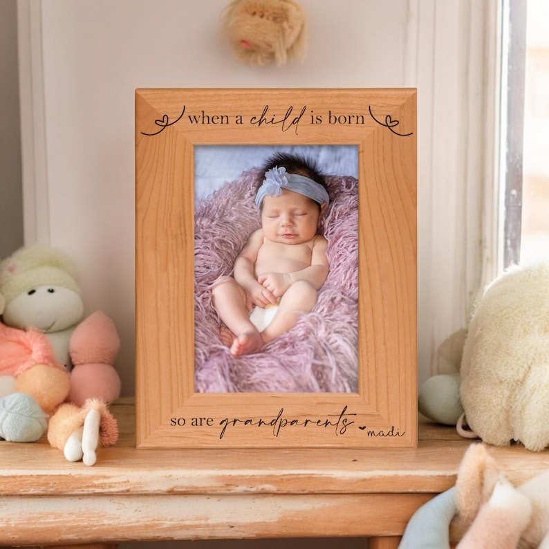 Pu&ograve; includere: Una cornice in legno marrone chiaro con la scritta "when a child is born so are grandparents". La cornice contiene una foto di un neonato con una fascia blu. Il bambino &egrave; sdraiato su una coperta viola.