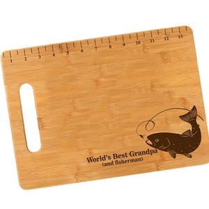 K&ouml;nnte beinhalten: Ein braunes Bambus-Schneidebrett mit einem aufgedruckten Lineal am oberen Rand. Das Brett hat einen Griff und ein lasergraviertes Design eines Fisches mit einer Angelschnur und dem Text "World's Best Grandpa (and fisherman)".