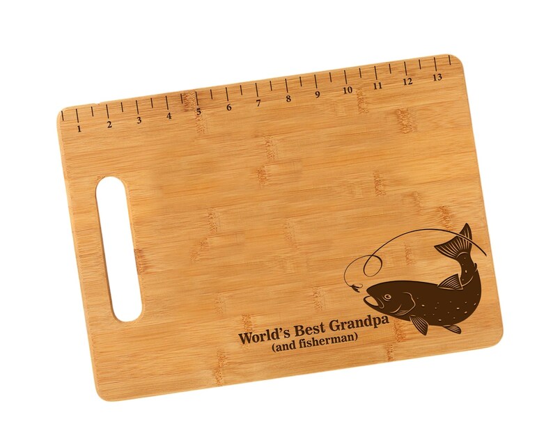 K&ouml;nnte beinhalten: Ein braunes Bambus-Schneidebrett mit einem aufgedruckten Lineal am oberen Rand. Das Brett hat einen Griff und ein lasergraviertes Design eines Fisches mit einer Angelschnur und dem Text "World's Best Grandpa (and fisherman)".