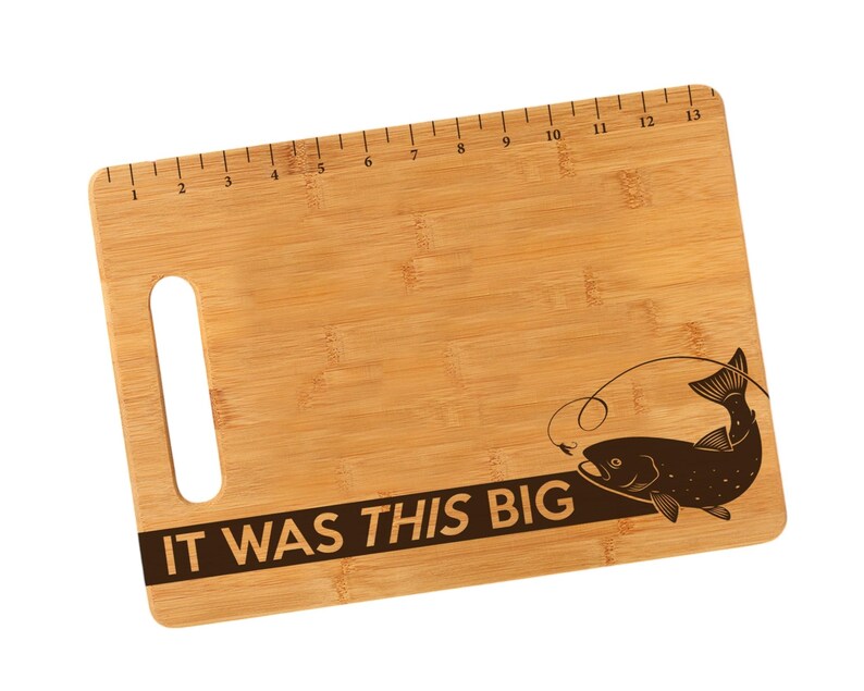 K&ouml;nnte beinhalten: Ein braunes Bambus-Schneidebrett mit einem Lineal, das auf der Oberkante aufgedruckt ist. Das Brett hat einen Griff und eine Fischsilhouette mit dem Text "IT WAS THIS BIG", der auf der Unterseite aufgedruckt ist.