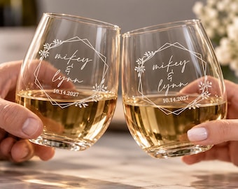 Può includere: Due calici da vino senza stelo trasparenti riempiti di vino bianco, ciascuno caratterizzato da un design a cornice geometrica bianca incisa, accenti floreali e il testo "mikey & lynn" seguito dalla data "14.10.2027" stampato sulla superficie.