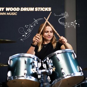 Puede incluir: Una mujer toca una bater&iacute;a con dos baquetas de madera. Las baquetas est&aacute;n etiquetadas con "5A Hickory Wood Drum Sticks" y "Make Your Own Music". El fondo es una pared gris oscura con el texto "Vital Signs Northwest" en blanco.