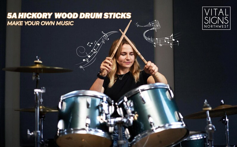 Puede incluir: Una mujer toca una bater&iacute;a con dos baquetas de madera. Las baquetas est&aacute;n etiquetadas con "5A Hickory Wood Drum Sticks" y "Make Your Own Music". El fondo es una pared gris oscura con el texto "Vital Signs Northwest" en blanco.