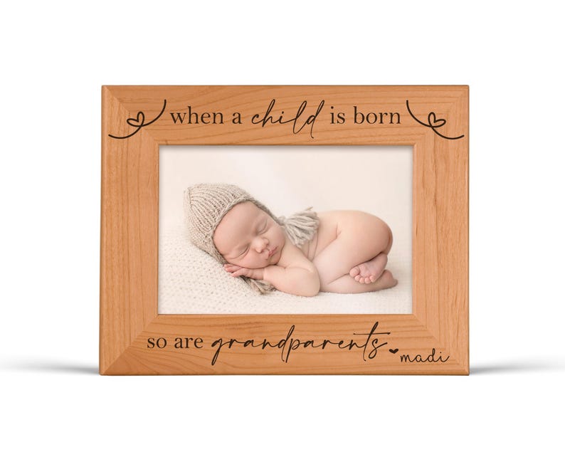 Pu&ograve; includere: Una cornice in legno marrone chiaro con la scritta "when a child is born so are grandparents" e una foto di un bambino. La cornice ha un design semplice. Il bambino indossa un cappello di lana e giace su una coperta bianca.
