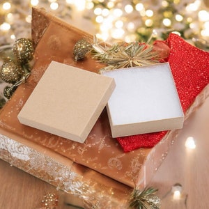 May include: Two gift boxes, one brown and one white, are sitting on a brown and gold patterned wrapping paper. The white box has a red ribbon tied around it. The boxes are surrounded by Christmas decorations, including gold ornaments and a red and gold garland.