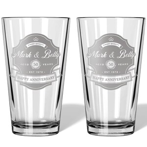 May include: Two clear glass pint glasses with a personalized design. The design features a vintage-style label with the text "Mark & Betty" and "Happy Anniversary". The label also includes the text "Aged 50 Years" and "Est 1972".