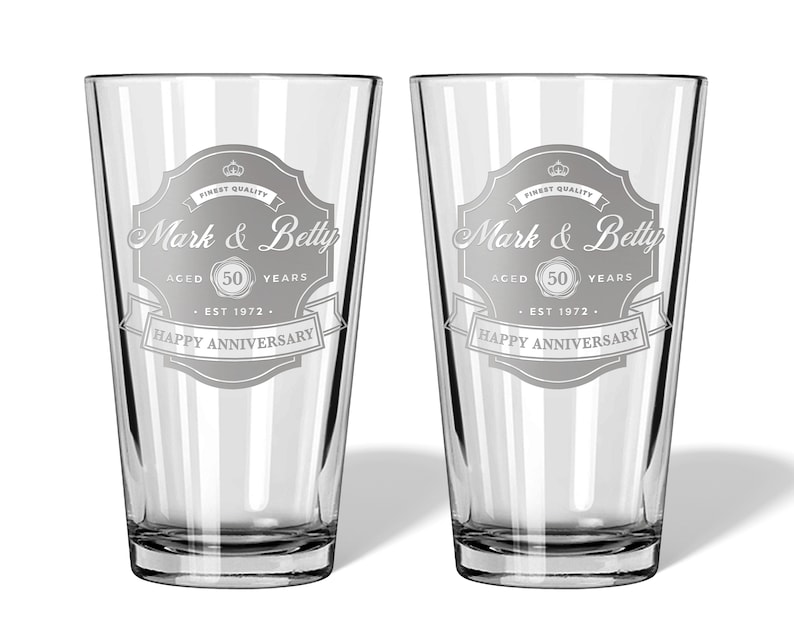 May include: Two clear glass pint glasses with a personalized design. The design features a vintage-style label with the text "Mark & Betty" and "Happy Anniversary". The label also includes the text "Aged 50 Years" and "Est 1972".