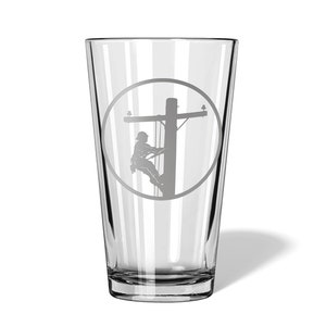 May include: A clear glass pint tumbler featuring a sandblasted circular design on the front. The etching depicts a utility worker wearing a hard hat and climbing a wooden utility pole with crossarms and insulators, rendered in a matte grey finish.