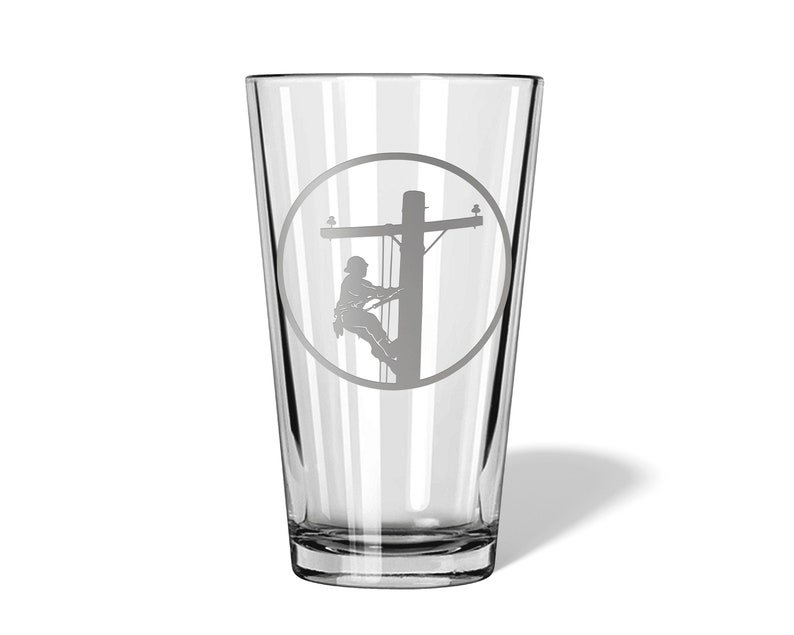 May include: A clear glass pint tumbler featuring a sandblasted circular design on the front. The etching depicts a utility worker wearing a hard hat and climbing a wooden utility pole with crossarms and insulators, rendered in a matte grey finish.