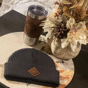 Puede incluir: Un gorro de punto negro con un parche de cuero marr&oacute;n que presenta un dise&ntilde;o de monta&ntilde;a. El gorro est&aacute; sobre una mesa junto a una taza de viaje de acero inoxidable con una envoltura de cuero marr&oacute;n y un dise&ntilde;o de monta&ntilde;a. La taza est&aacute; sobre un mantel individual redondo tejido con un dise&ntilde;o floral.