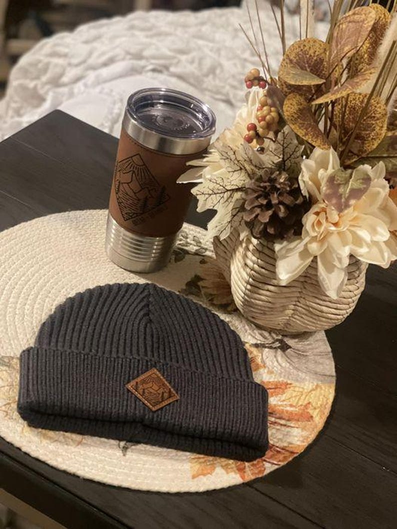 Puede incluir: Un gorro de punto negro con un parche de cuero marr&oacute;n que presenta un dise&ntilde;o de monta&ntilde;a. El gorro est&aacute; sobre una mesa junto a una taza de viaje de acero inoxidable con una envoltura de cuero marr&oacute;n y un dise&ntilde;o de monta&ntilde;a. La taza est&aacute; sobre un mantel individual redondo tejido con un dise&ntilde;o floral.