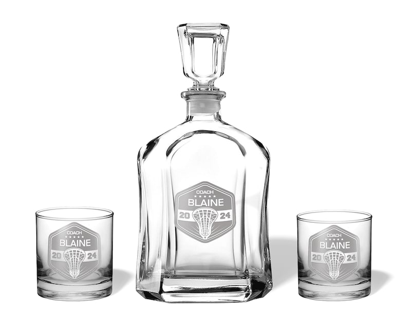 May include: A clear glass decanter set with two matching lowball glasses. The decanter has a square stopper. Each piece is etched with a lacrosse graphic and the text "COACH BLAINE 2024".