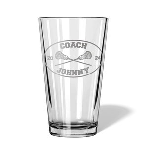 May include: A clear pint glass with a custom design. The design features the text "COACH JOHNNY" inside an oval, with crossed lacrosse sticks and the numbers "20" and "24". The glass is empty and has a shadow.