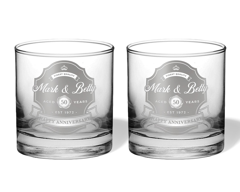 May include: Two clear glass tumblers with a silver etched design. The design features a banner with the text "Mark & Betty" and "Happy Anniversary". The banner also includes the text "Aged 50 Years" and "Est 1972".