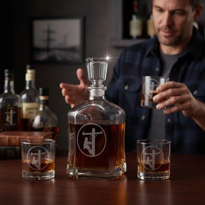 May include: A clear glass decanter and two matching lowball glasses, each featuring an etched circular design of a utility worker on a power pole. The decanter has a square base and a faceted glass stopper, all filled with an amber-coloured liquid.