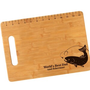 K&ouml;nnte beinhalten: Ein braunes Bambus-Schneidebrett mit einem aufgedruckten Lineal am oberen Rand. Das Brett hat einen ausgeschnittenen Griff und ein lasergraviertes Design eines Fisches mit dem Text "World's Best Dad (and fisherman)".