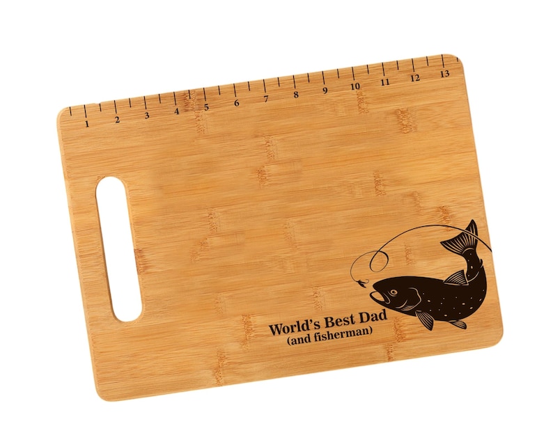K&ouml;nnte beinhalten: Ein braunes Bambus-Schneidebrett mit einem aufgedruckten Lineal am oberen Rand. Das Brett hat einen ausgeschnittenen Griff und ein lasergraviertes Design eines Fisches mit dem Text "World's Best Dad (and fisherman)".