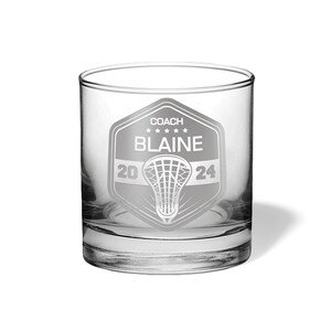 May include: A clear glass tumbler with a personalized lacrosse design. The design includes the text "COACH BLAINE" above a lacrosse net graphic, with "2024" below. The glass is cylindrical with a thick base and is suitable for beverages.