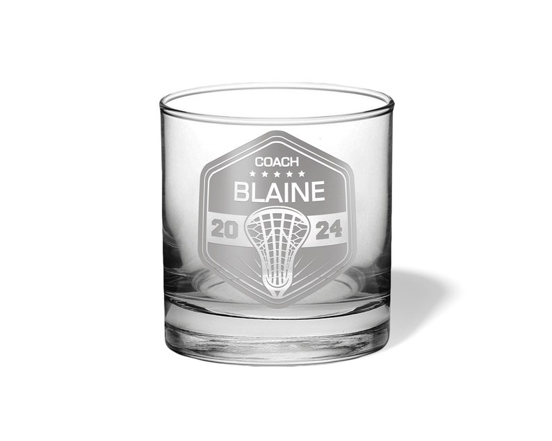 May include: A clear glass tumbler with a personalized lacrosse design. The design includes the text "COACH BLAINE" above a lacrosse net graphic, with "2024" below. The glass is cylindrical with a thick base and is suitable for beverages.