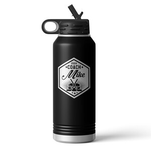 May include: A matte black insulated water bottle with a silver base and a black lid featuring a straw. The front displays a silver hexagonal emblem with the text 2024 COACH Mike, accompanied by a graphic of a hockey puck and two crossed sticks.