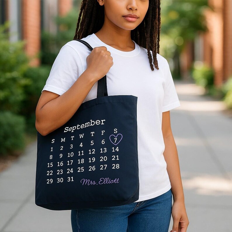 May include: A navy blue tote bag displaying a September calendar, with the 7th date marked with a heart. The bag also features the text "September" and "Mrs. Elliott". The bag is held by a person wearing a white t-shirt and blue jeans.