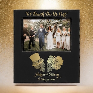 May include: Black leather picture frame with a gold inscription that reads "Til Death Do Us Part". The frame has a photo of a wedding party and a gold engraved design of two skeletons with flowers. The inscription below the skeletons reads "Aaron & Stacey October 31, 2023".